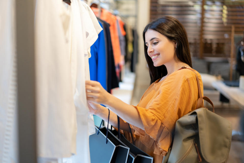 A woman with long dark hair wearing a casual top and carrying a backpack shops for clothes, holding several shopping bags