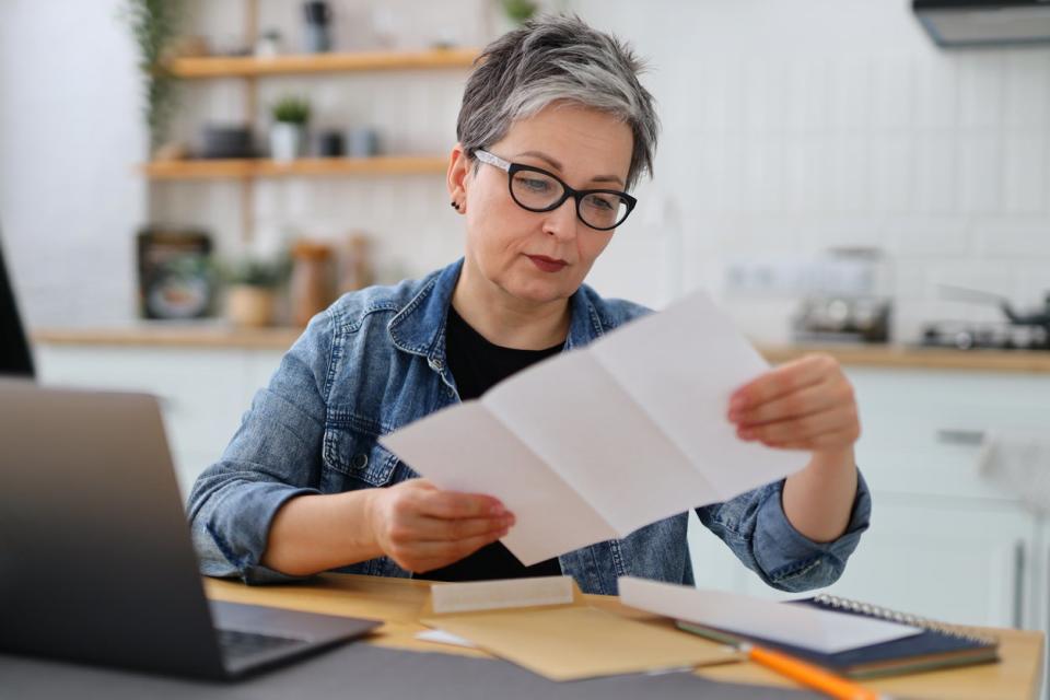 A woman inspects a piece of paper while sitting in front of a laptop in the kitchen.