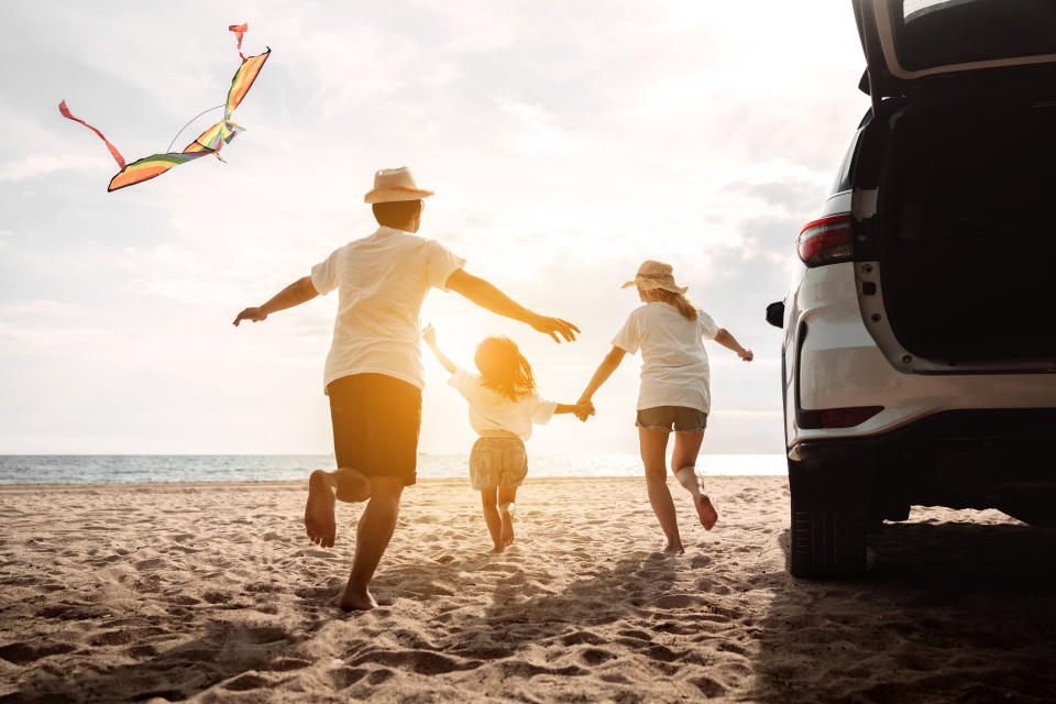 Two adults and a child run on a beach away from a parked SUV with the rear hatch open toward a low-flying kite and the sun-strewn horizon.