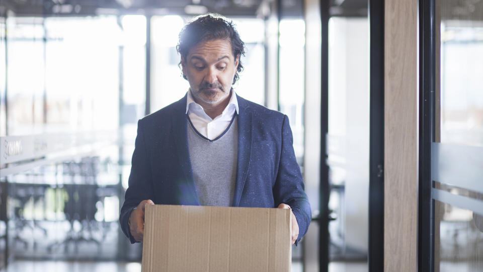 Latin executive man of approximately 50 years dressed in blue suit gray coat and white shirt walks through the corridors of the company where he works with a cardboard box where he carries his personal things in his hands which looks sadly since it was fired from his work half body picture.