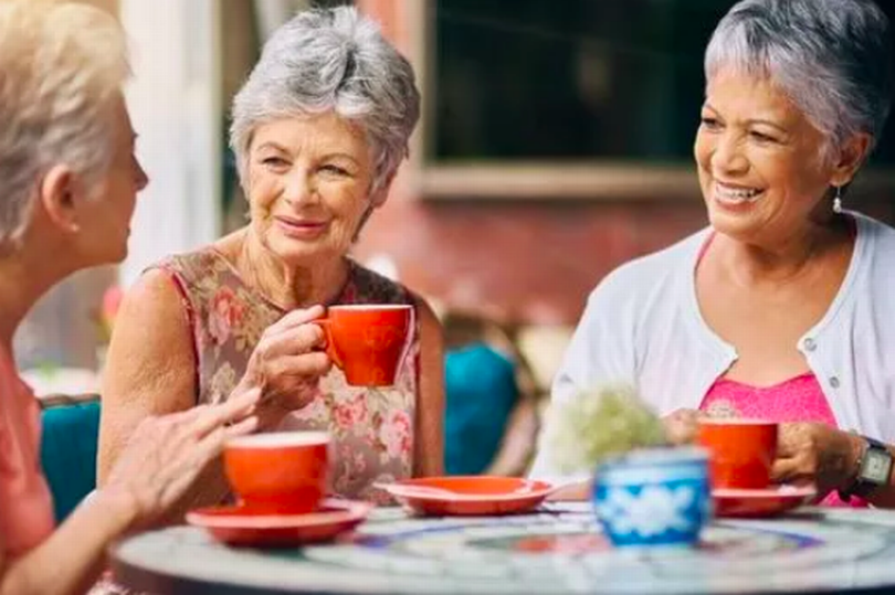 A trio of older women are sitting at a table smiling and drinking coffee