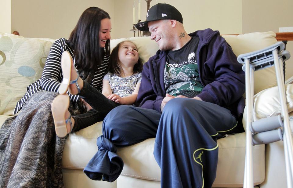 a woman, child, and man look at each other and smile on a white coach, with a crutch leaning against the couch next to the man