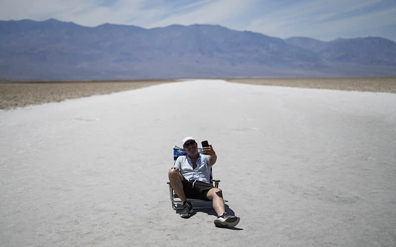 A man sits in a low chair as he takes a selfie on the salt flats at Badwater Basin in Death Valley National Park. The white ground that his chair is on is seen extending away into the distance, where is also see a faraway mountain range.