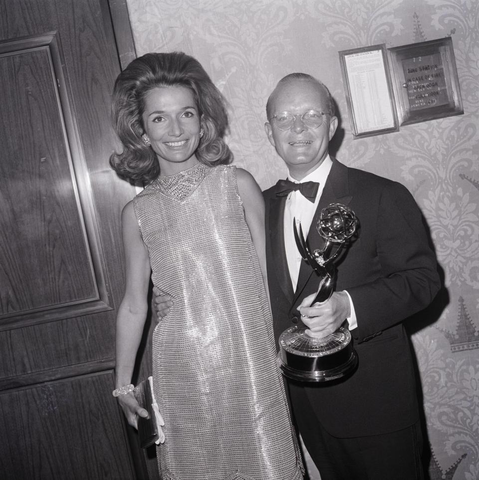 lee radziwill and truman capote stand next to each other and smile for a photo while standing in front of a wall with a door, he holds an emmy award and wears a dark suit with a bowtie, she wears a sleeveless dress