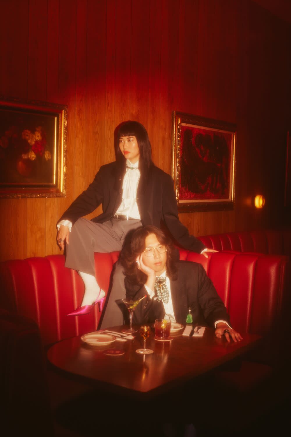 An Asian couple sit at restaurant booth with drinks, each looking off to the side. They are wearing vintage suits.