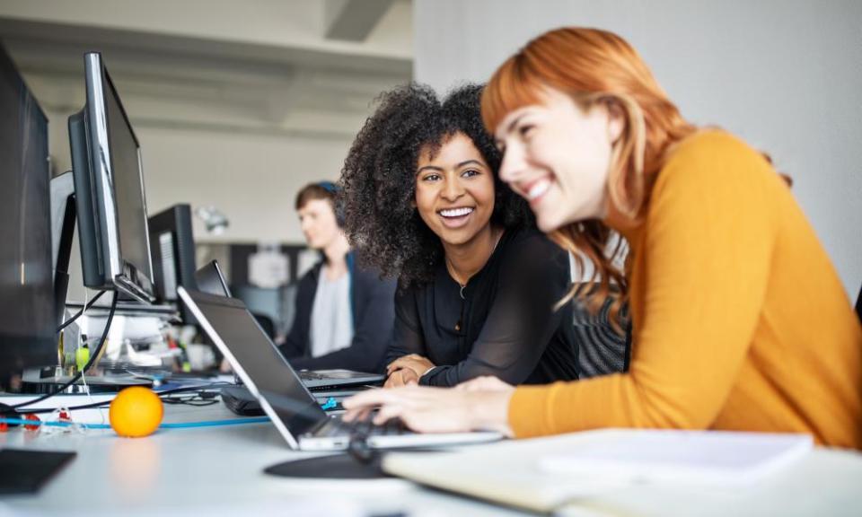 Two young women working together on laptop with male colleague in background.