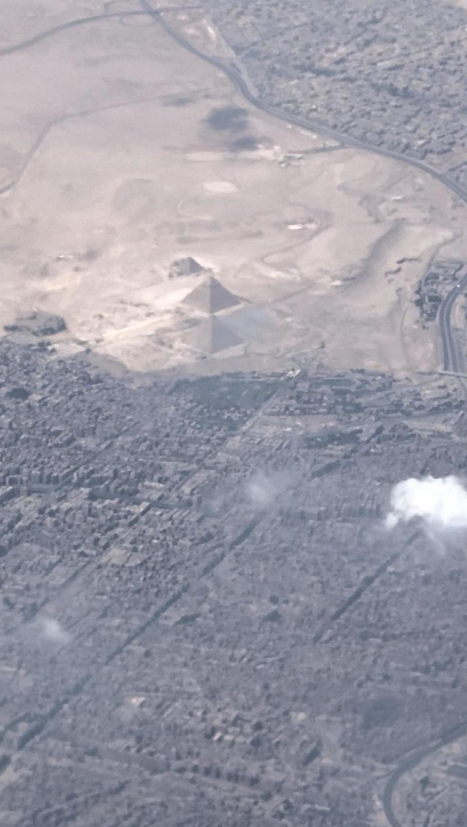 Aerial view of the Great Pyramid of Giza surrounded by the city of Giza, with visible roads and sparse clouds. No people are present in the image