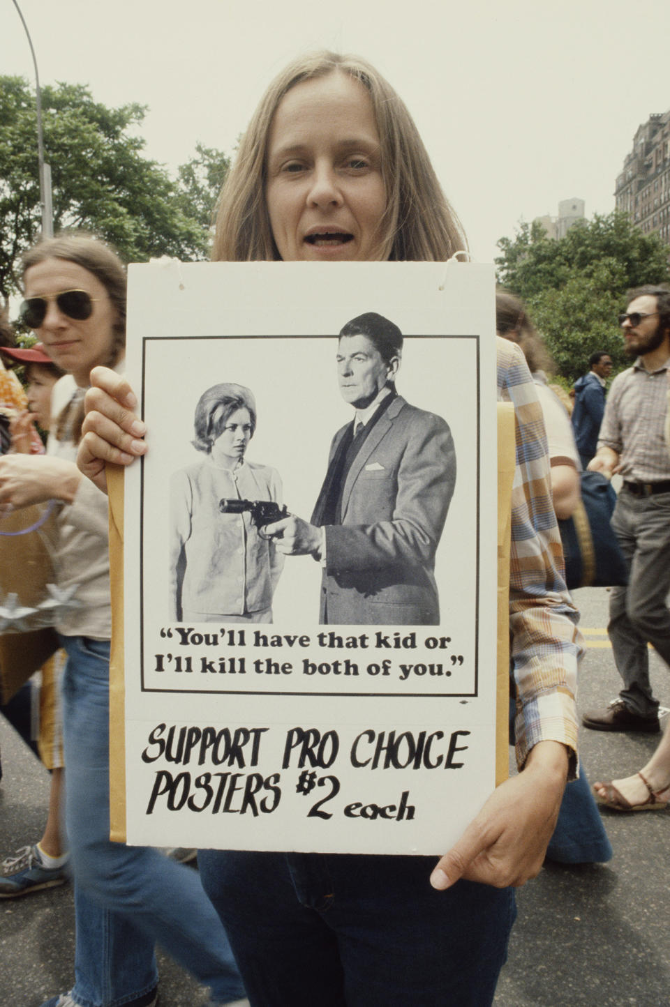 A demonstrator's sign shows Ronald Reagan holding up a gun with the text "You'll have that kid or I'll kill the both of you"