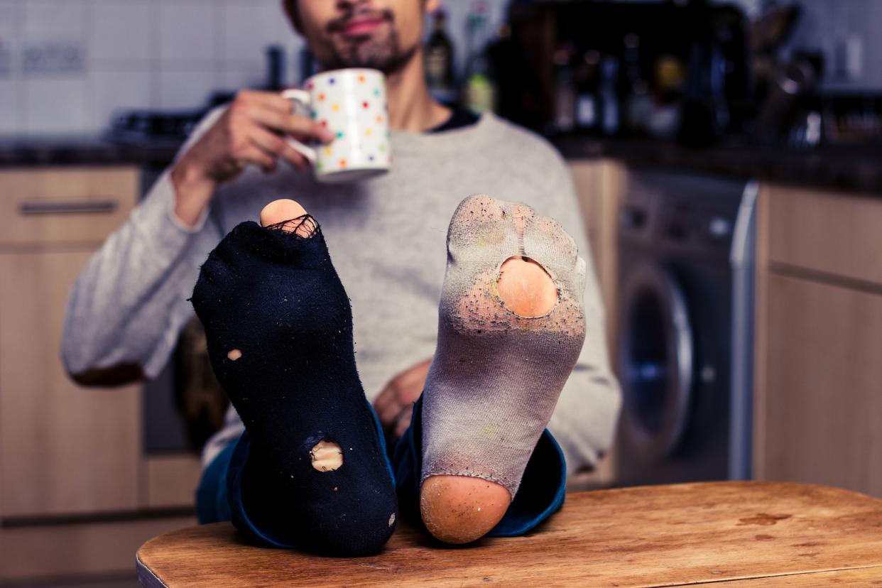 young man with worn out socks, one white, one black, is resting his feet on a table in his kitchen while relaxing with a cup of coffee