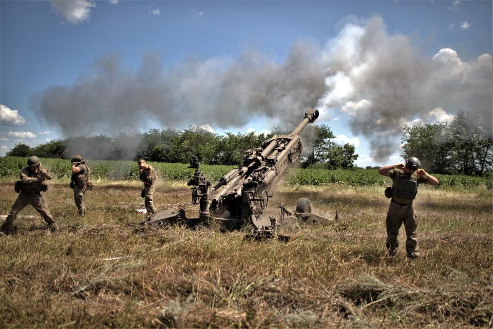 Russian troops covering their ears as a howitzer is fired in a field, producing smoke.