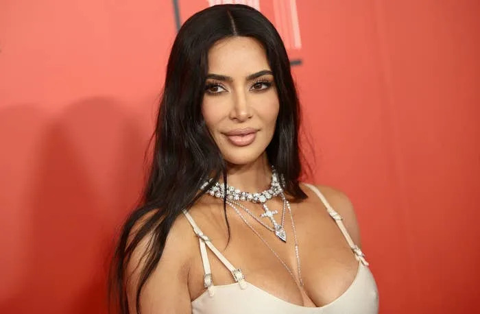 Kim Kardashian smiles slightly, her long hair down. She is wearing a neutral-colored dress with thin straps and multiple layered necklaces