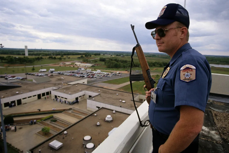 A guard with a baseball cap and rifle stands high above a prison compound, looking out at low buildings, parked cars and a flat landscape that extends to a cloudy sky.