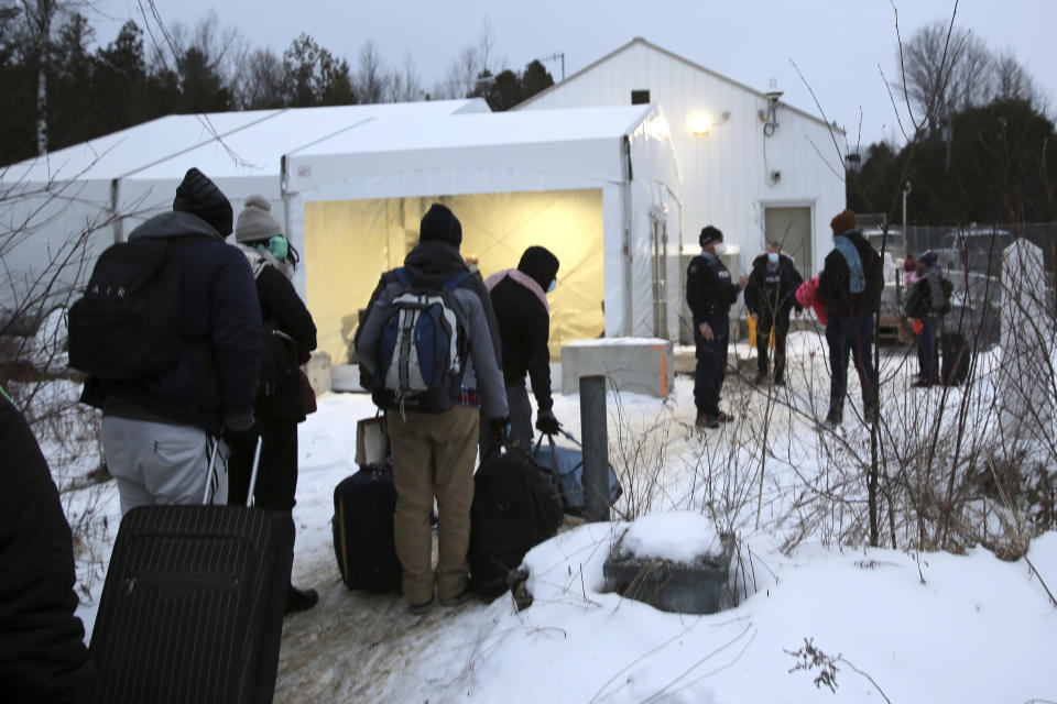 Migrants line up on the border of the United States, foreground, and Canada, background, at a reception center for irregular borders crossers, in Saint-Bernard-de-Lacolle, Quebec, Canada, Wednesday Jan. 12, 2022, in a photo taken from Champlain, N.Y. They are crossing the U.S.-Canadian border into Saint-Bernard-de-Lacolle, Quebec, where they are arrested by the Royal Canadian Mounted Police and then allowed to make asylum claims. The process was halted for most cases after the 2020 outbreak of COVID-19, but the Canadian government changed its policy in November, allowing the process to continue. (AP Photo/Wilson Ring)