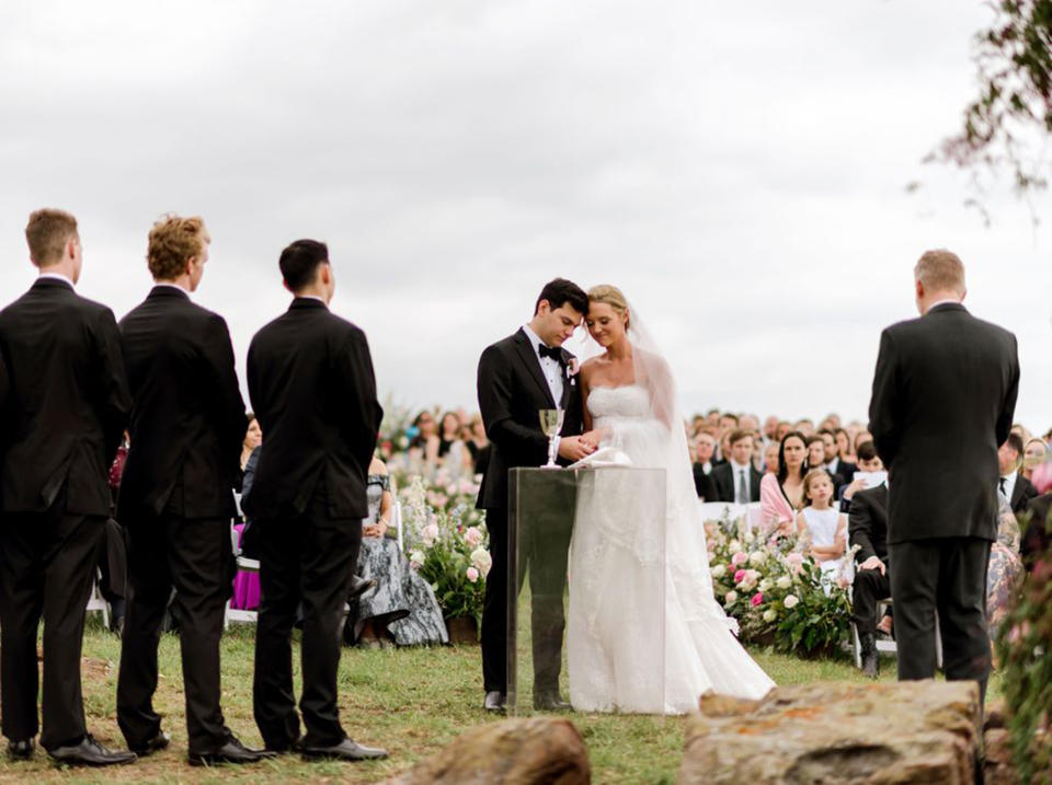 A bride and groom stand side by side in front of a crowd of family and friends outside under an overcast sky (Courtesy of Ashley Alexander Photography)