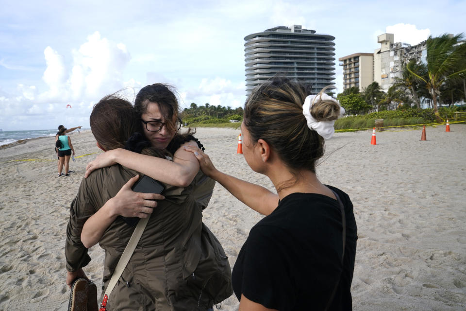 Faydah Bushnaq, of Sterling, Va., center, is hugged by Maria Fernanda Martinez, of Boca Raton, Fla., as they stand outside of a 12-story beachfront condo building which partially collapsed, Friday, June 25, 2021, in the Surfside area of Miami. Bushnaq is vacationing and stopped to write "Pray for their Souls" in the sand. (AP Photo/Lynne Sladky)