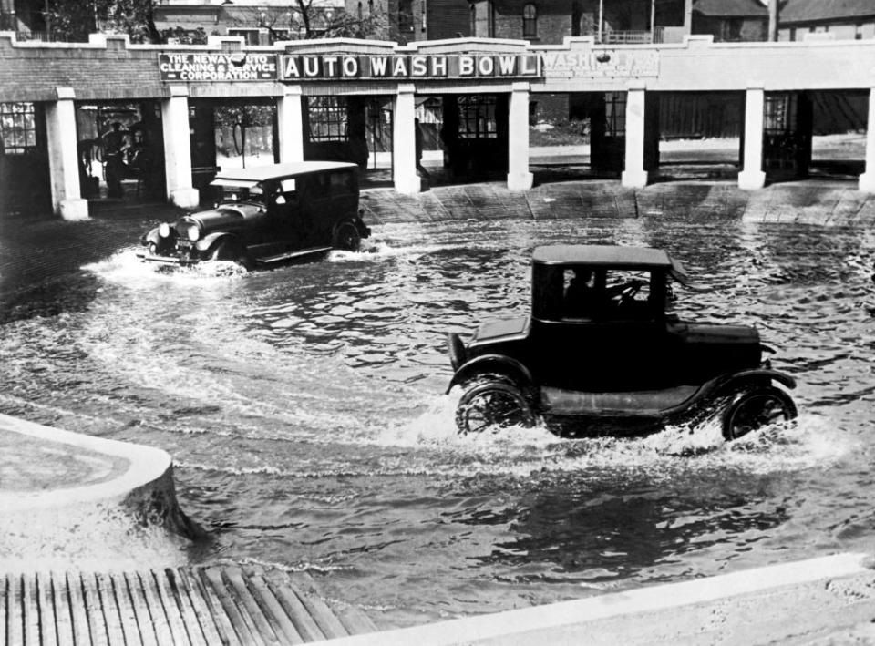 Vintage cars driving through circular water-filled channels at the Auto Wash Bowl, an early car wash facility featuring automatic water jets