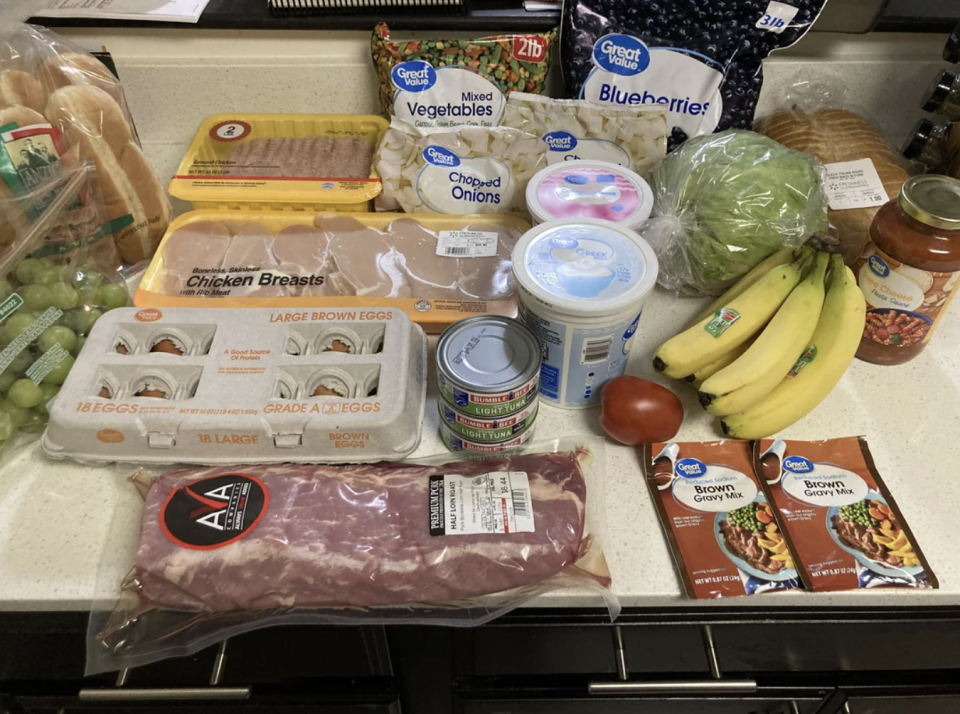 A kitchen counter with a variety of groceries including eggs, chicken, bananas, canned tuna, blueberries, grapes, vegetables, chopped onions, pork ribs, and spice mix