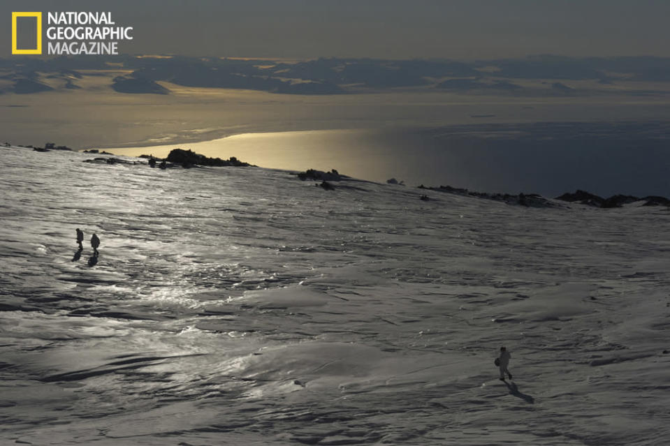 An unearthly landscape. On the upper slopes of Mount Erebus, the ice has been polished and textured by the wind. In the distance, sea ice, open ocean, and the mountains and dry valleys of the Antarctic mainland. Three figures—tiny in this vast place—prepare to return to camp after a cold day's work. (photo © Carsten Peter/National Geographic)