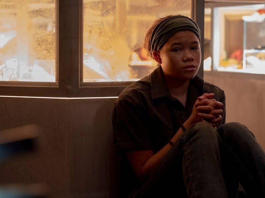 riley in the last of us. she's forlorn and sitting with her back to a display case, her hands clasped in front of her. she has blonde hair styled in braids, with a headband over her hairline.