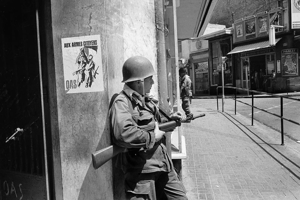 FILE - In this May 15, 1962 file photo, a French soldier guards a street corner in Oran, Algeria. French President Emmanuel Macron announced a decision to speed up the declassification of secret documents related to Algeria's seven-year war of independence from 1954 to 1962. On the wall is a poster of the nationalist Secret Armed Organization, calling for citizens to take up arms against Algerian independence. (AP Photo/Horst Faas, File)