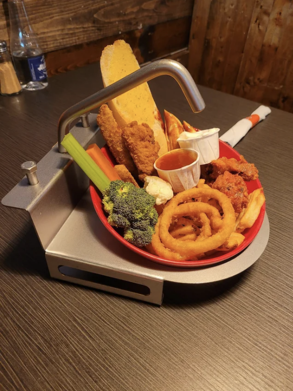 A platter of assorted appetizers including onion rings, fried chicken strips, vegetables, and dipping sauces, served creatively with a faucet-like decoration