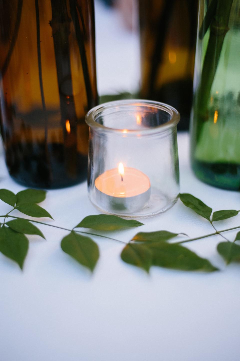 A candle sits in a jar on a table with leaves in front of it.