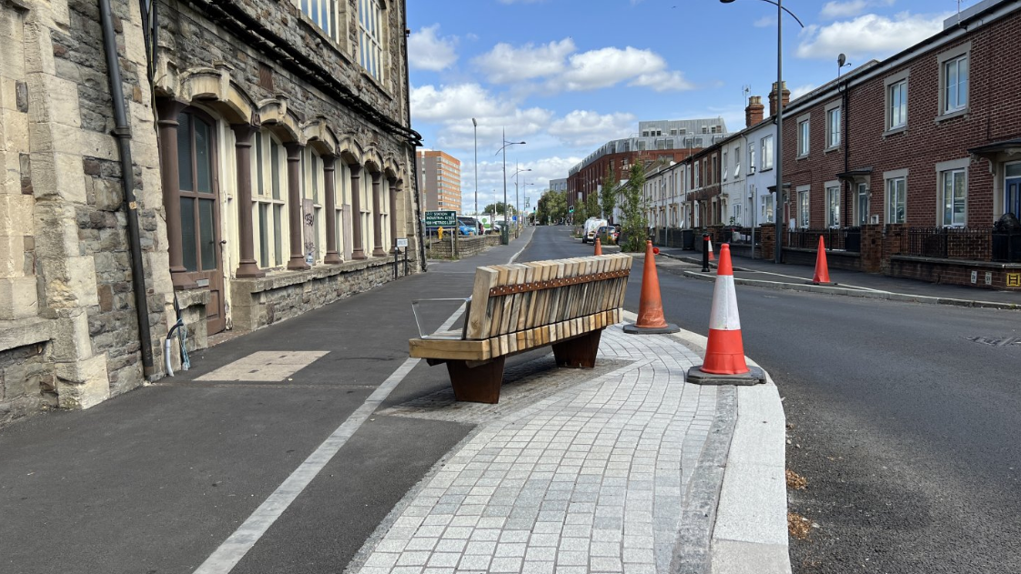 A wooden bench on a street faces a wall at an unlikely angle