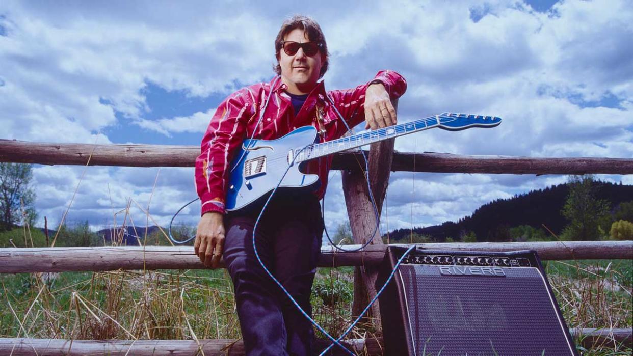  Steve Miller leaning on a fence holding a guitar, with an amplifier at his feet. 