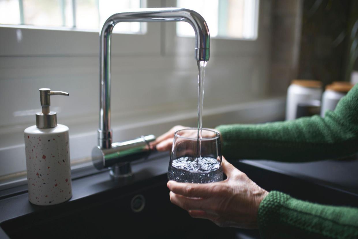 Hand pouring a glass of water from a tap, over a sink, soap dispenser pictured beside it, can see persons arms in green wool jumper