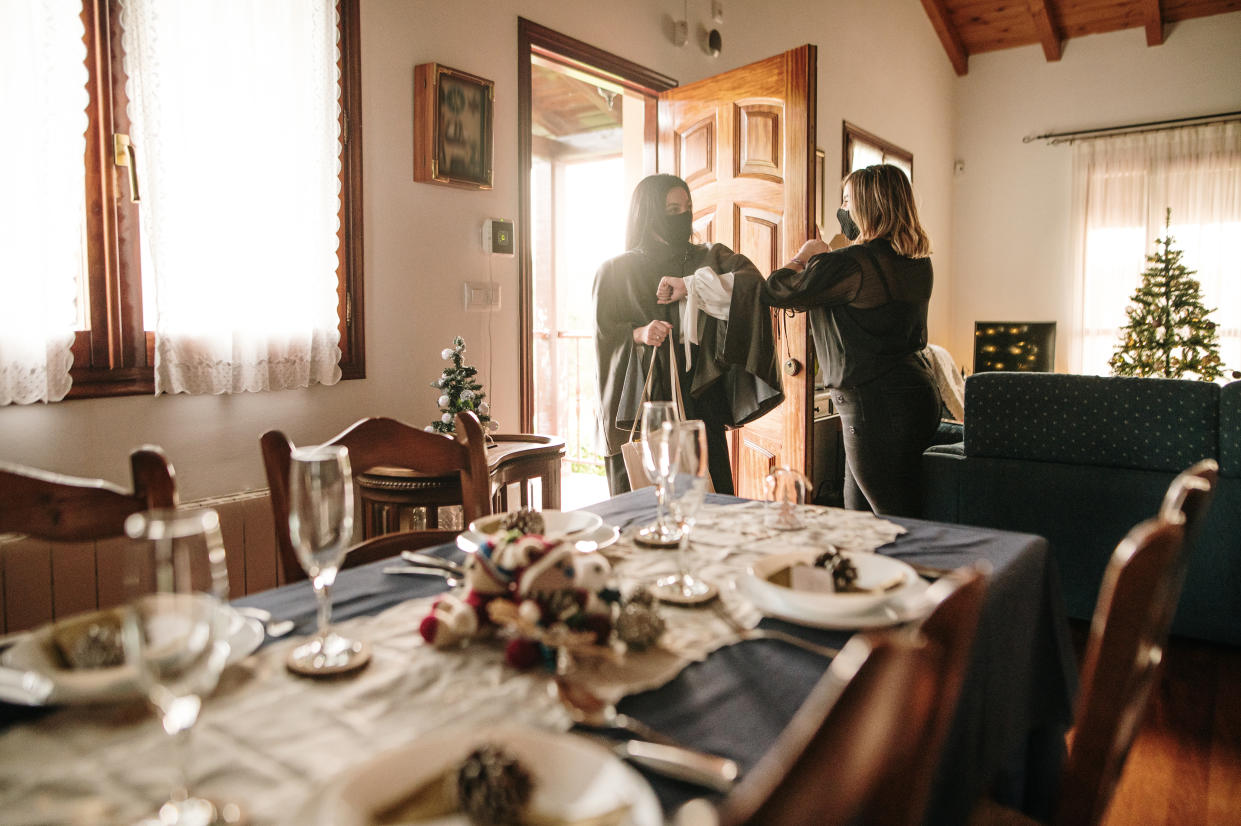 Two masked women greet each other at the entrance of a house, with a Christmas-decorated dinner table.