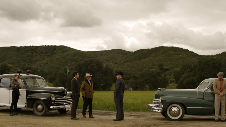 five men stand on a country road with green mountains in the background and a cloudy gray sky above.