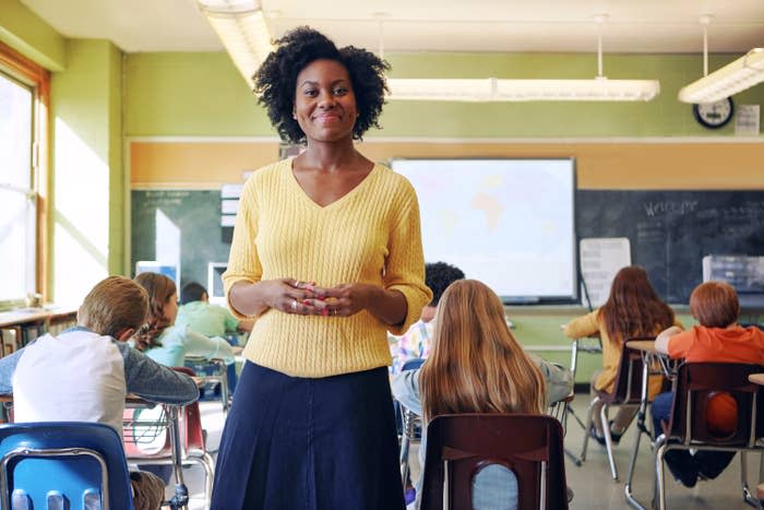 a teacher smiling at the camera in her classroom while her students work at their desks