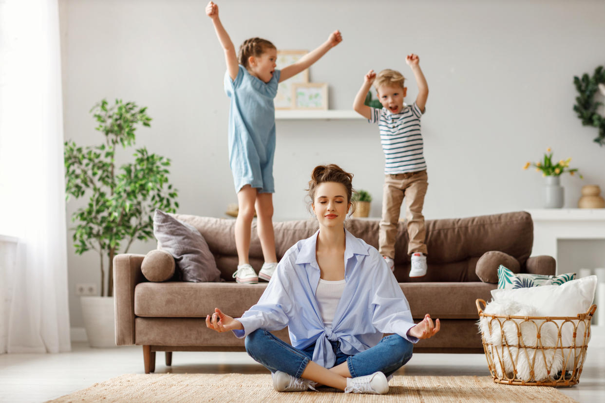 Happy mother with closed eyes meditating in lotus pose on floor trying to save inner harmony while excited children jumping on sofa and screaming in light spacious living room