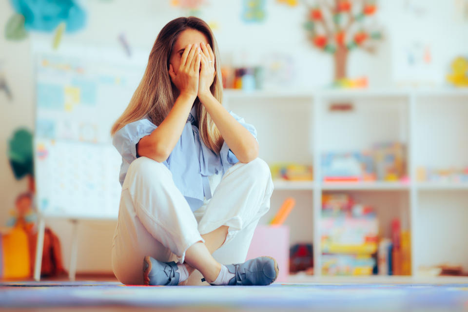 Woman sitting on floor with hands covering her face, in a room with shelves and children's items