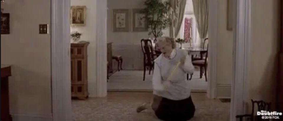 Robin Williams, dressed as Mrs. Doubtfire in a domestic setting, is energetically dancing with a broom