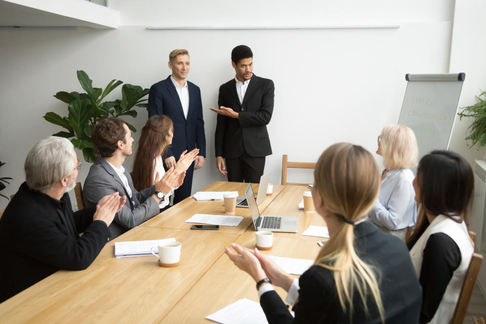 Man in suit pointing to another man in suit while table of professionals applauds.