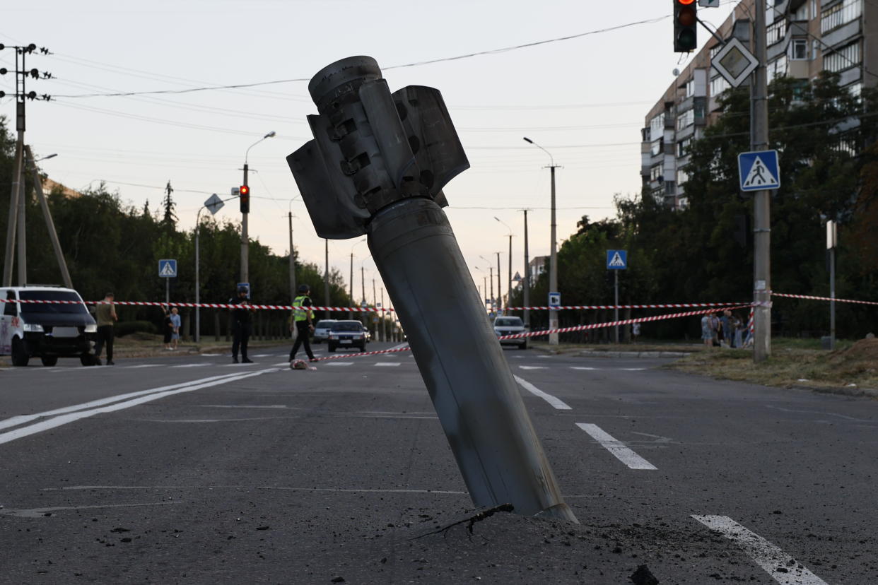 A intact missile is half embedded into the middle of a road, in an area taped off, while police stand by, presumably a safe distance away.