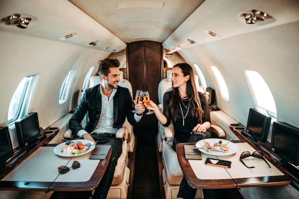 Rich couple making a toast with champagne glasses while eating canapes aboard a private jet.