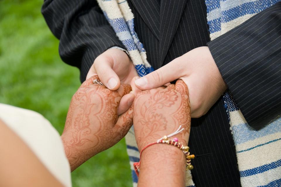 A photo of two people holding hands, with the woman's hands decorated in brownish red designs.