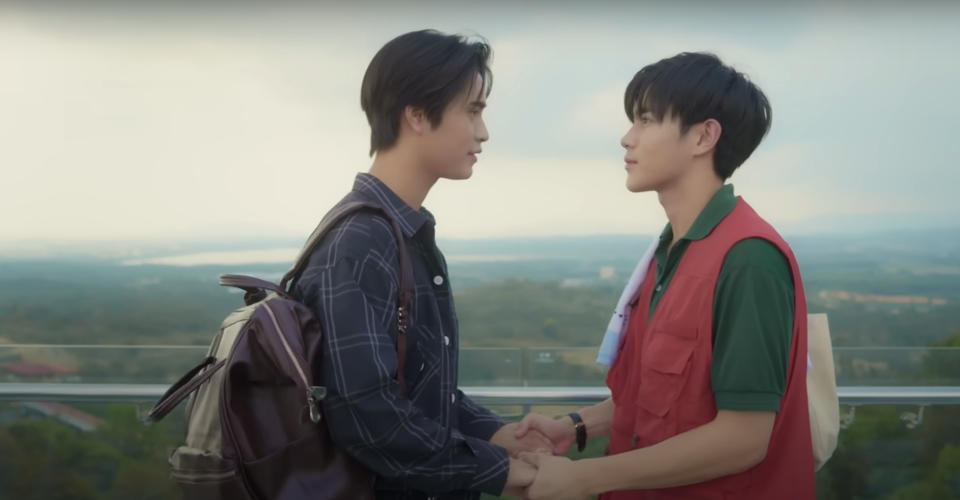 Two individuals, one in a plaid shirt with a backpack and another in a red vest, hold hands and gaze at each other on an outdoor terrace