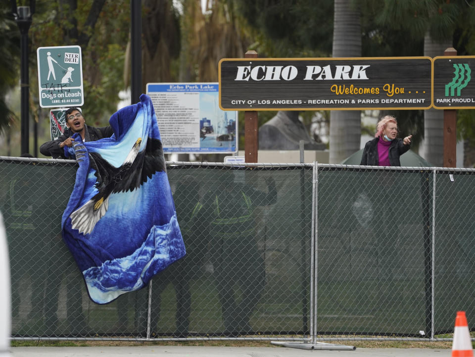 A homeless activist waves a blanket over a fence as Los Angeles City sanitation workers move inside the closed perimeter of Echo Park Lake in Los Angeles, Thursday, March 25, 2021. A newly installed fence surrounded the park Thursday after a late-night confrontation between police and vocal demonstrators who oppose the city's effort to remove a large homeless encampment and perform extensive repairs of the site. People who were already in tents at Echo Park Lake were allowed to remain overnight but were given 24-hour notice to leave, Police Chief Michel Moore said in a social media post. (AP Photo/Damian Dovarganes)