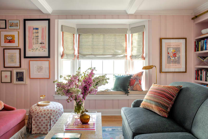 Window seat with cushion and throw pillows at bay window with Roman shades in pink library with gallery wall.