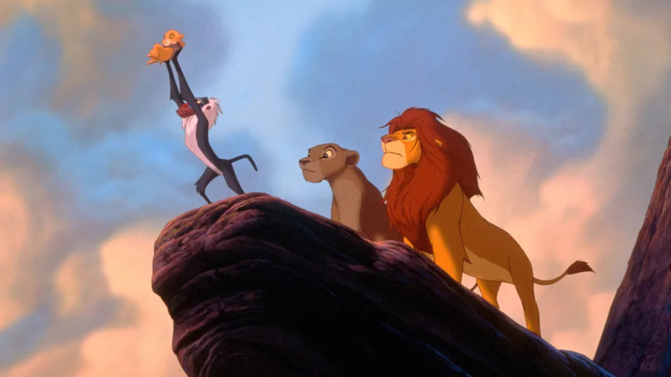Rafiki holds Simba up into the air on Pride Rock, with Mufasa and Nala behind him. 