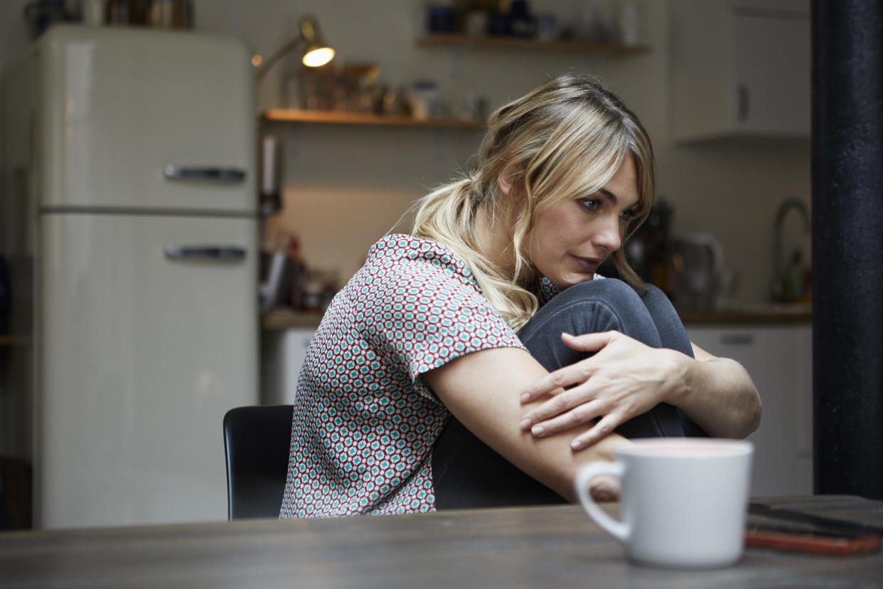A woman sits at a kitchen table, hugging her knees to her chest.
