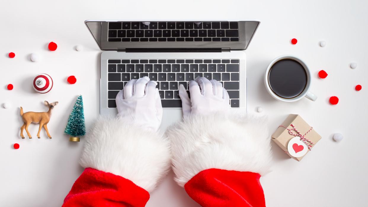  Santa hands in white gloves with red sleeves and white fluffy cuffs seen from above, typing on a laptop, surrounded by small Christmas ornaments. 