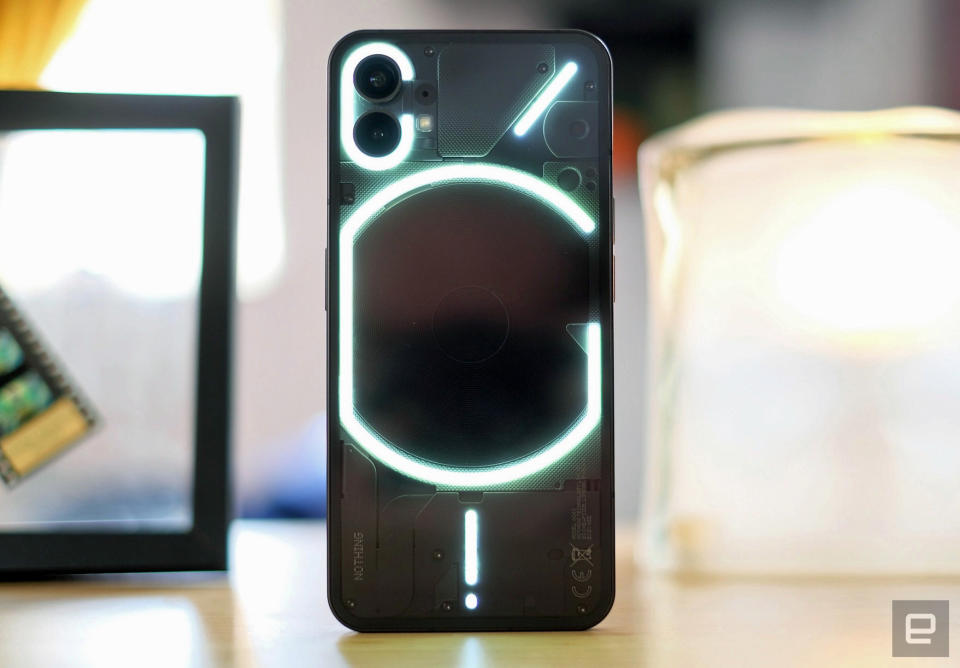 The new Nothing Phone 1 stands on its end on a table. The phone is black with a see-through back showing a glowing series of lines and rings.
