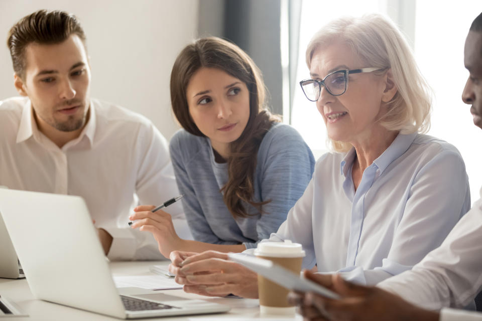 Focused young students interns workers making notes listening to old senior aged female manager coach mentor leader teacher talking at group office meeting instructing business work team with laptop