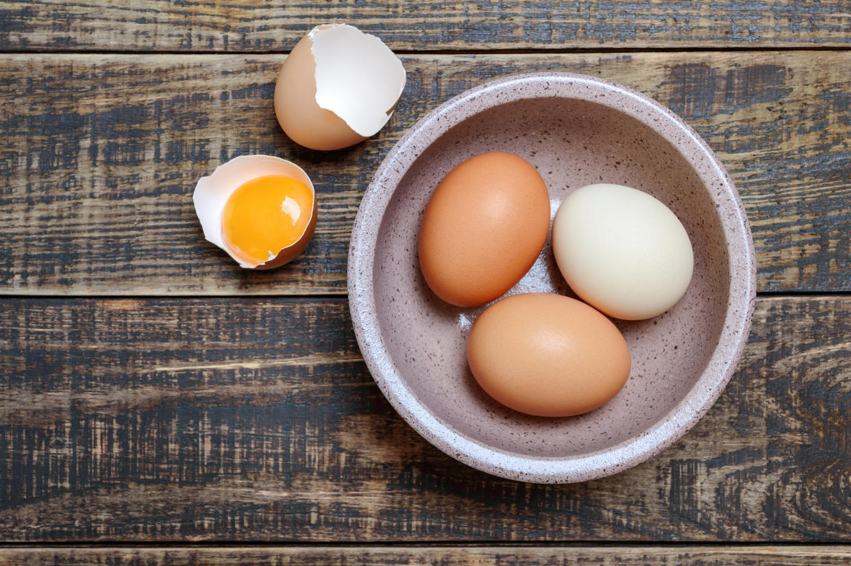 A broken egg next to a bowl containing three eggs in their shells.