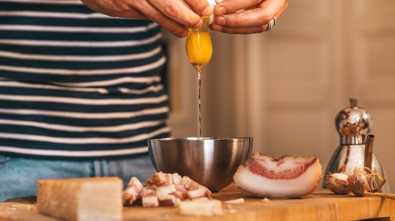 Person breaking an egg into a metal bowl on a cutting board next to cured pork and a block of parmesan