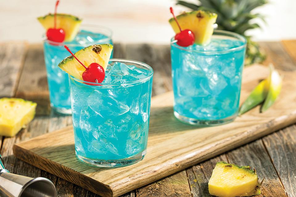 Three tropical blue-hued cocktails garnished with cherries and pineapple slices